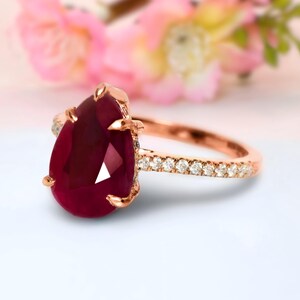 Puede incluir: Un anillo de oro rosa con un gran rubí rojo oscuro en forma de pera. La banda está adornada con pequeños diamantes transparentes. El anillo se presenta sobre un fondo floral borroso.