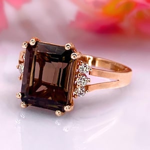 Könnte beinhalten: Ein roségoldener Ring mit einem großen, rechteckigen, rauchbraunen Edelstein. Der Ring ist mit kleinen, klaren Edelsteinen und dekorativen Golddetails verziert. Das Band teilt sich in der Nähe des Edelsteins in zwei Teile.