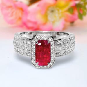 Op de afbeelding: Een zilveren ring met een grote, rechthoekige, dieprode edelsteen in het midden, omringd door een halo van kleinere, heldere stenen. De band is ook bezet met meerdere rijen kleinere, heldere stenen. De achtergrond is een zachte vervaging van roze en groen.