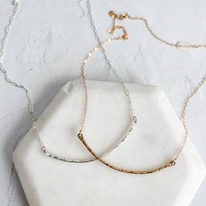 May include: Two delicate necklaces with a curved bar pendant. One necklace is silver and the other is gold.