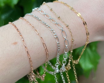 May include: A set of five delicate chain bracelets in gold, silver, and rose gold. The bracelets are layered on a person's wrist, showcasing different chain styles and colors.