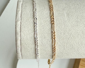 May include: Two Figaro chain bracelets, one silver-toned and one gold-toned, displayed side-by-side. The bracelets have a classic design with alternating elongated and round links. The chains are shown against a neutral, light-colored background.