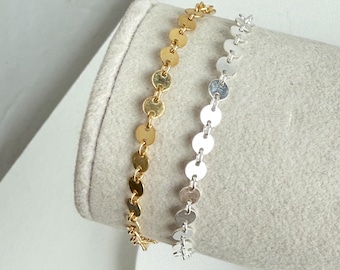 May include: Two delicate bracelets, one gold-toned and one silver-toned, each featuring a chain of small, round, flat discs. The bracelets are displayed on a neutral-colored, cylindrical form, showcasing their design and texture. The gold bracelet is on the left, and the silver bracelet is on the right.