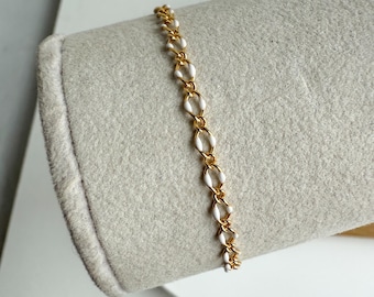 May include: A gold-colored bracelet with white oval-shaped links. The bracelet is displayed on a light gray surface. The jewelry piece is delicate and elegant, suitable for everyday wear or special occasions.