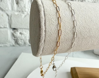 May include: Two delicate chain necklaces, one gold-toned and one silver-toned, with elongated oval links. The necklaces are displayed on a light gray cylindrical jewelry display, showcasing their design and texture. The background is a white brick wall.