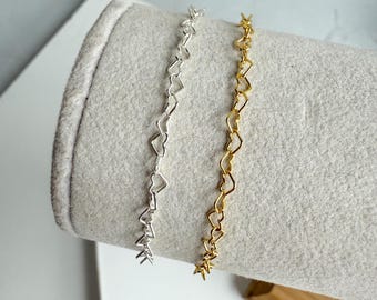 May include: Two delicate bracelets, one silver and one gold, each featuring a repeating heart design. The bracelets are displayed on a light gray, felt-covered form, showcasing their intricate link patterns. The gold bracelet has a warm, shiny finish.