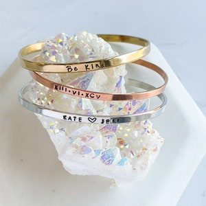 May include: Three gold, silver, and copper bracelets stacked on top of each other. The bracelets are engraved with the words "Be Kind", "XIII.VI.XCIV", and "KATE ♥ JEFF".