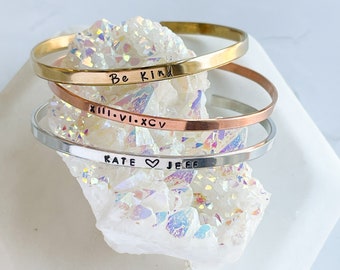 May include: Three gold, silver, and copper bracelets stacked on top of each other. The bracelets are engraved with the words "Be Kind", "XIII.VI.XCIV", and "KATE ♥ JEFF".