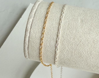 May include: Two delicate chain bracelets, one gold-toned and one silver-toned, are displayed on a soft, off-white surface. The bracelets feature an oval link design. The gold bracelet is on the left, and the silver bracelet is on the right.