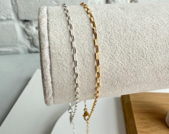 May include: Two delicate chain necklaces, one silver and one gold, displayed on a gray fabric. The silver chain has a rectangular link design, while the gold chain has a similar design. Both necklaces are shown with their clasps.
