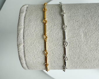 May include: Two delicate chain bracelets, one gold-toned and the other silver-toned, are displayed on a gray, textured surface. Each bracelet features a series of elongated links connected by small, circular rings, creating a minimalist design.