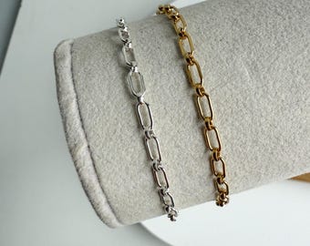 May include: Two chain link bracelets, one silver and one gold, displayed on a gray surface. The bracelets have an elongated oval link design. The gold bracelet is on the right, and the silver bracelet is on the left.