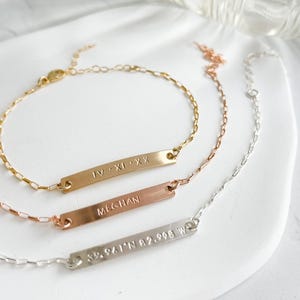 May include: Three gold, rose gold, and silver chain bracelets with personalized bar charms. The gold charm reads "IV.XI.XX", the rose gold charm reads "MEGHAN", and the silver charm reads "89.941°N 82.998°W".