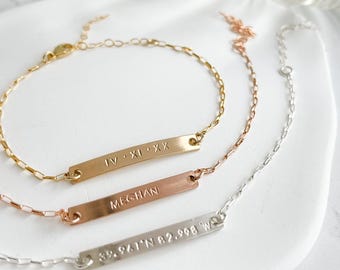 May include: Three gold, rose gold, and silver chain bracelets with personalized bar charms. The gold charm reads "IV.XI.XX", the rose gold charm reads "MEGHAN", and the silver charm reads "89.941°N 82.998°W".