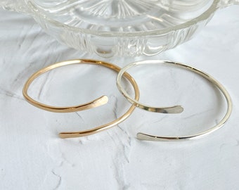 May include: Two simple, minimalist bracelets, one gold and one silver, with a hammered finish. The bracelets are open and have a slightly curved shape.