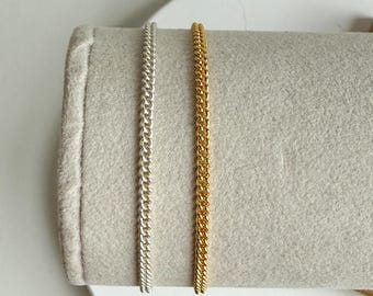 May include: Two curb chain necklaces, one silver and one gold, displayed on a gray felt surface. The chains have a classic, interlocking link design. The gold chain has a bright, shiny finish, while the silver chain has a polished look.