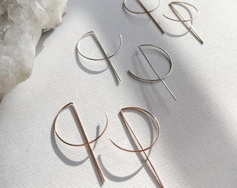 May include: Three pairs of geometric hoop earrings in silver, gold, and rose gold. Each earring features a half-circle shape with a straight line extending from the bottom.