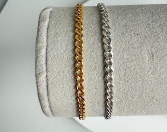 May include: Two curb chain bracelets, one gold-toned and one silver-toned, displayed side-by-side. The chains have a classic, interlocking link design. The bracelets are presented on a light gray, cylindrical display.