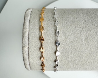 May include: Two delicate chain bracelets, one gold-toned and one silver-toned, each featuring a series of small, starburst-shaped charms. The bracelets are displayed on a light gray surface, showcasing their intricate design and metallic sheen.