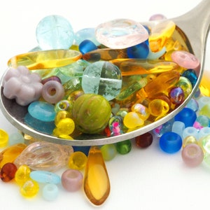 May include: A stainless steel spoon filled with a vibrant mix of glass beads. The beads are in diverse shapes and sizes, including round, teardrop, and flower-shaped, in shades of blue, yellow, green, and pink.