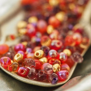 May include: A close-up of a variety of colourful glass beads in shades of red, pink, purple, and gold. The beads are arranged in a loose pile on a silver surface.