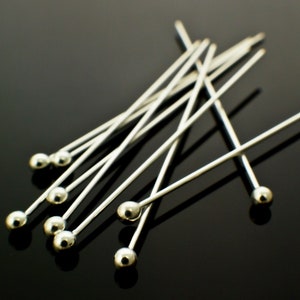 May include: A group of silver-toned straight pins with round heads. The pins are arranged in a random pattern on a black surface.
