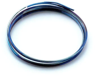 Puede incluir: Una longitud de alambre metálico fino enrollado que presenta un acabado azul iridiscente vibrante con sutiles tonos púrpuras y plateados. El alambre está enrollado en forma circular, mostrando una superficie lisa y reflectante en toda su extensión.