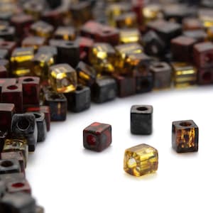 Puede incluir: Un montón grande de pequeñas cuentas cuadradas de vidrio en tonos de marrón, rojo y dorado. Cuatro cuentas están separadas del montón, mostrando sus colores y texturas individuales.