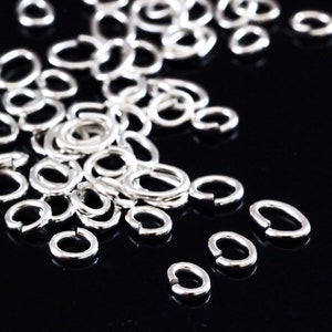 May include: A pile of silver jump rings, some are scattered on a black surface. The jump rings are oval shaped and have a smooth finish.