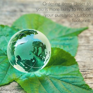 May include: A glass globe representing the Earth sits on a green leaf. The globe is clear and shows the continents in green. The text "Ordering items closer to you is more likely to reduce your purchase's carbon footprint." is above the globe.