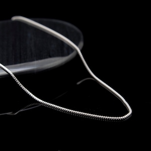 May include: A silver chain necklace with a box chain design. The chain is lying on a black surface.