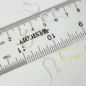 Puede incluir: Varios ganchos para pendientes de pl&aacute;stico transparente y curvado est&aacute;n esparcidos alrededor de una regla Westcott. La regla muestra marcas en pulgadas y cent&iacute;metros, con los ganchos midiendo aproximadamente 2 cm de largo.