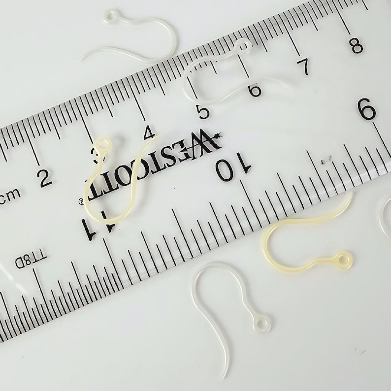 Puede incluir: Varios ganchos para pendientes de pl&aacute;stico transparente y curvado est&aacute;n esparcidos alrededor de una regla Westcott. La regla muestra marcas en pulgadas y cent&iacute;metros, con los ganchos midiendo aproximadamente 2 cm de largo.