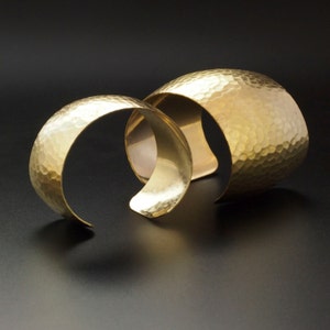 以下が含まれることがあります： 金色のハンマーで叩いた金属製のブレスレットが2つ。ブレスレットは重ねられています。両方のブレスレットはハンマーで叩いた金属製で、質感があります。