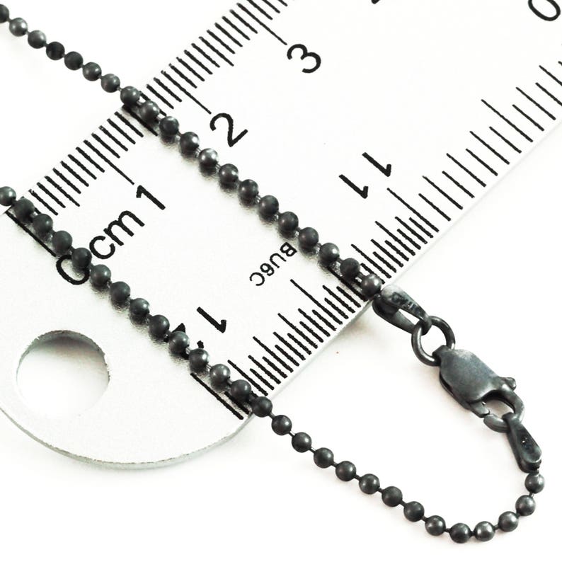 May include: A black ball chain necklace with a lobster clasp. The chain is measured against a ruler, showing it is 17 inches long.