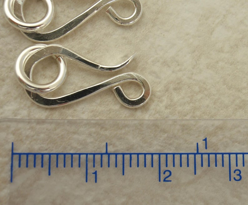 K&ouml;nnte beinhalten: Zwei silberfarbene Metallverschl&uuml;sse, jeder mit einer Schlaufe und einem gebogenen Haken, werden vor einem hellbraunen Hintergrund gezeigt. Ein Lineal ist unter den Verschl&uuml;ssen platziert und zeigt, dass sie etwa 2,5 cm lang sind.