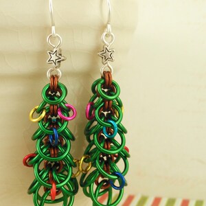 Puede incluir: Un par de pendientes de árbol de Navidad de color verde, plateado y multicolor. Los pendientes están hechos de anillos de metal y tienen un encanto de estrella en la parte superior.