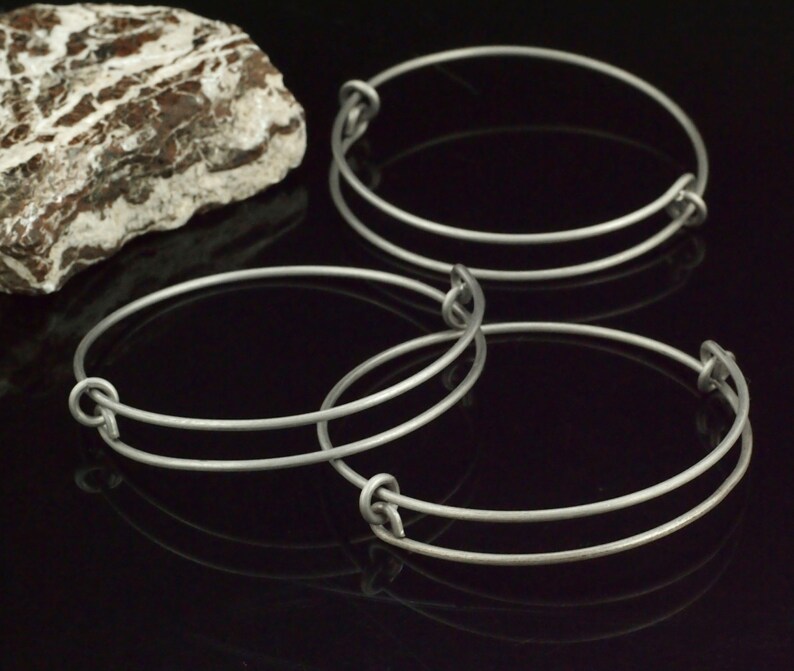 Puede incluir: Varios brazaletes ajustables de color plateado con un dise&ntilde;o de nudo. Los brazaletes est&aacute;n hechos de metal fino y tienen un estilo simple y minimalista. Una roca texturizada est&aacute; en el fondo.