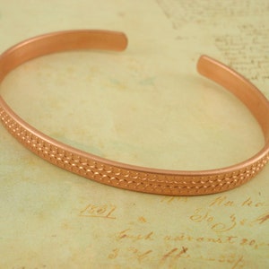 May include: A copper bangle bracelet with a textured, patterned design. The bracelet is a simple, minimalist style.