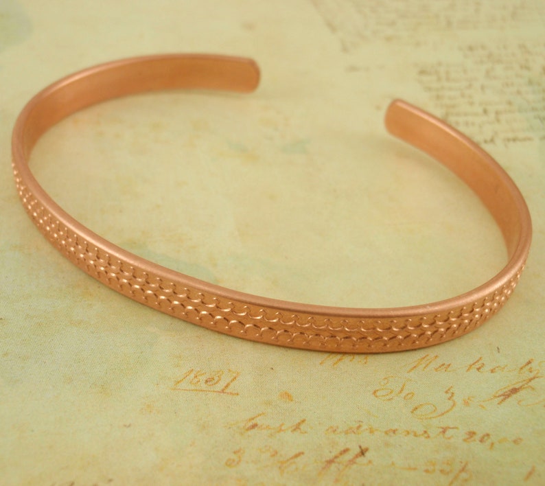 May include: A copper bangle bracelet with a textured, patterned design. The bracelet is a simple, minimalist style.