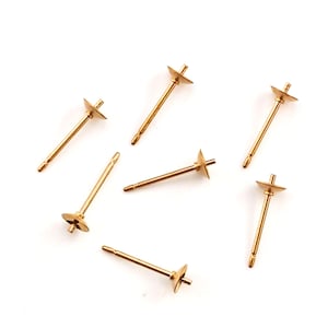 May include: A collection of six gold-toned metal earring posts, each featuring a thin, straight pin and a small, flat, circular cup at the top designed for attaching decorative elements. The components are arranged in a scattered, loose formation.