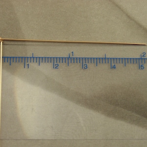 Puede incluir: Dos alfileres de cabeza de metal dorado fino colocados contra una regla transparente. El alfiler horizontal mide aproximadamente 5 cm de largo, mientras que el vertical se extiende hacia abajo, ambos con una peque&ntilde;a cabeza circular plana.