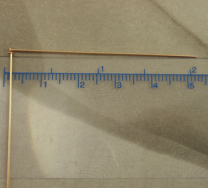 Puede incluir: Dos alfileres de cabeza de metal dorado fino colocados contra una regla transparente. El alfiler horizontal mide aproximadamente 5 cm de largo, mientras que el vertical se extiende hacia abajo, ambos con una peque&ntilde;a cabeza circular plana.