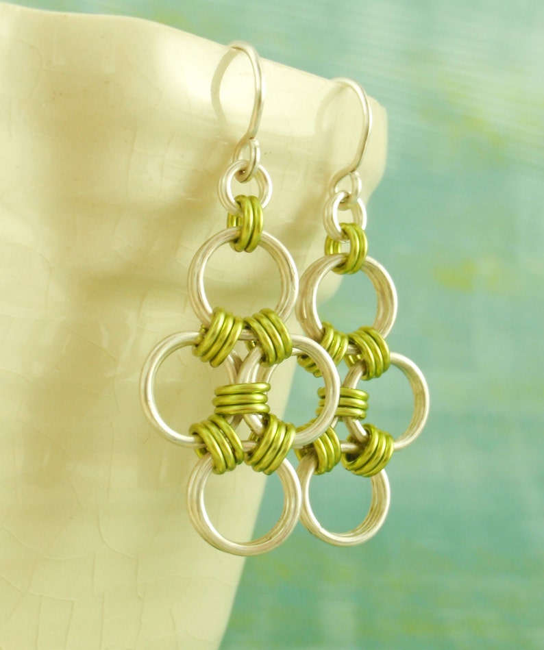 May include: Silver earrings with a green chainmail design. The earrings are made of metal and have a dangling design.