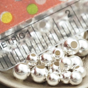 May include: A close-up of a pile of silver-coloured round beads. The beads are smooth and shiny, and they are arranged in a random pattern. The beads are approximately 1.25 cm in diameter.