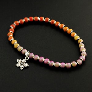 May include: A beaded bracelet featuring a gradient of spherical beads transitioning from orange to yellow to purple, separated by small silver-toned spacers. A silver-toned metal flower charm hangs from the bracelet, which is arranged in a circle.