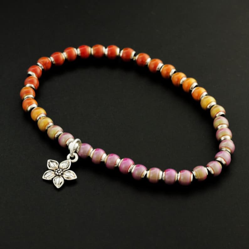 May include: A beaded bracelet featuring a gradient of spherical beads transitioning from orange to yellow to purple, separated by small silver-toned spacers. A silver-toned metal flower charm hangs from the bracelet, which is arranged in a circle.