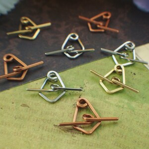 May include: A collection of handmade metal jewelry findings, including diamond-shaped clasps and straight bar closures. The clasps are in various metallic finishes: gold, silver, and copper. The jewelry components are arranged on a textured, green and brown surface.