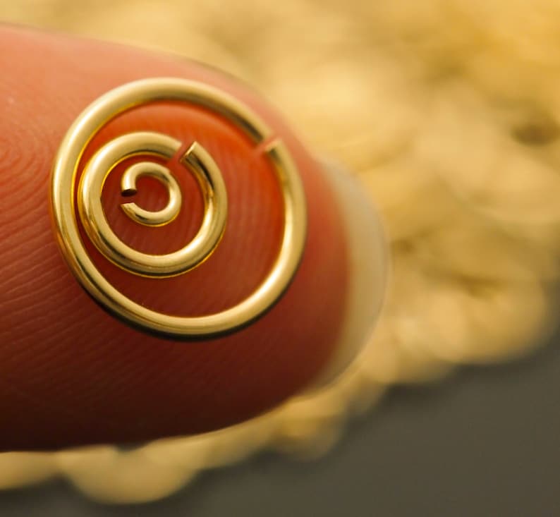 Op de afbeelding: Een close-up van een kleine, goudkleurige spiraalvormige oorbel. De oorbel heeft een eenvoudig, minimalistisch ontwerp met een cirkelvorm. De oorbel wordt door een vinger vastgehouden en de achtergrond is wazig.
