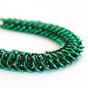 May include: A green and black chainmail bracelet made of metal rings. The bracelet is made with a pattern of alternating green and black rings.
